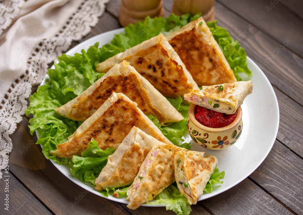 Sliced ham and cheese wraps. Fried patties or mini pies made from thin Armenian lavash served with tomato sauce on a white plate with lettuce leaves.