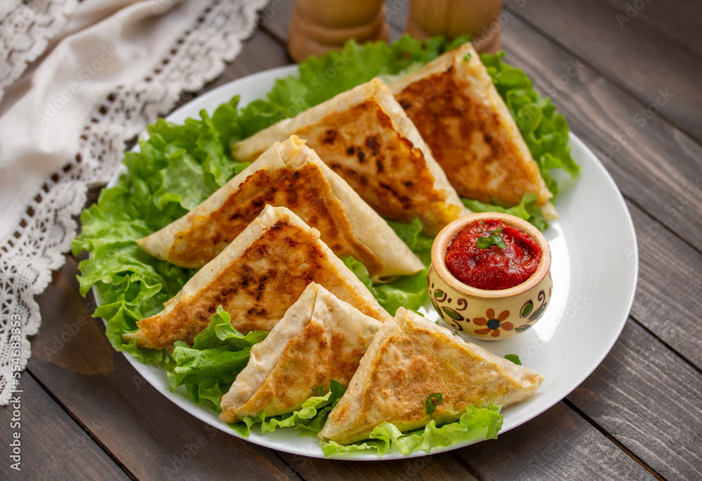 Mini Flatbread Wraps with cheese and ham filling. Fried patties from thin Armenian lavash served with tomato sauce on a white plate with lettuce leaves. Selective focus, horizontal, wooden table. Mini Flatbread Wraps with cheese and ham filling. Fried patties from thin Armenian lavash served with tomato sauce on a white plate with lettuce leaves. Selective focus, horizontal, wooden table.