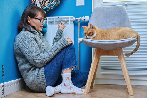 Woman in knitted woolen sweater with pet cat warming near heating radiator