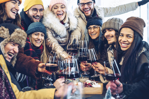 Happy multiracial friends toasting red wine at restaurant pub patio - Group of young people wearing winter clothes having fun at outdoors winebar table - Dining life style and friendship concept