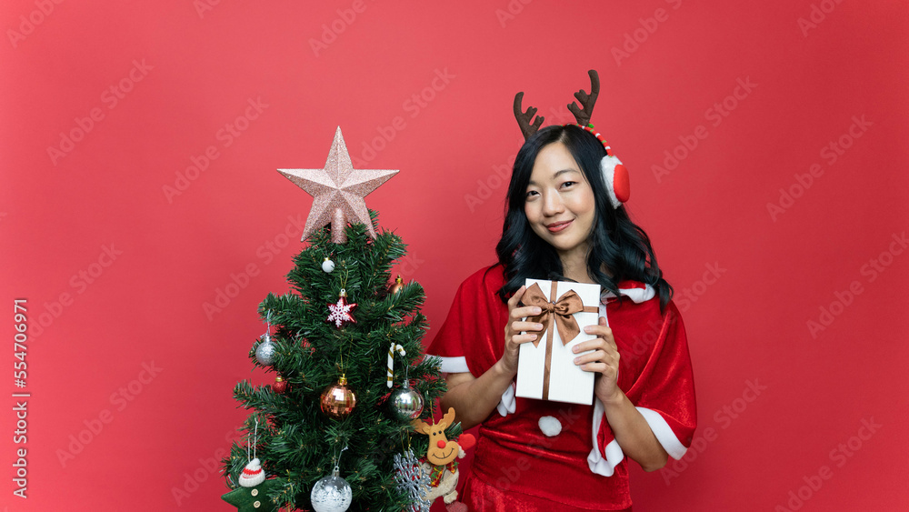 Young beautiful woman with red santy costume holding gift box. The scene has a Christmas tree and a red background. Young beautiful woman with red santy costume holding gift box. The scene has a Christmas tree and a red background.