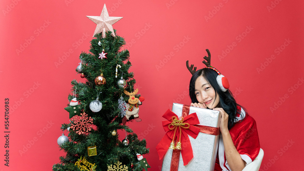 Young lady with red santy costume holding big gift box. The scene has a Christmas tree and a red background. Young lady with red santy costume holding big gift box. The scene has a Christmas tree and a red background.