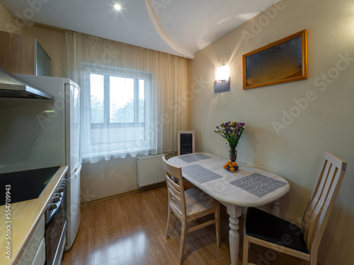 Modern interior of kitchen in apartment. Table and chairs. Fridge, oven and fan. Window with tulle.