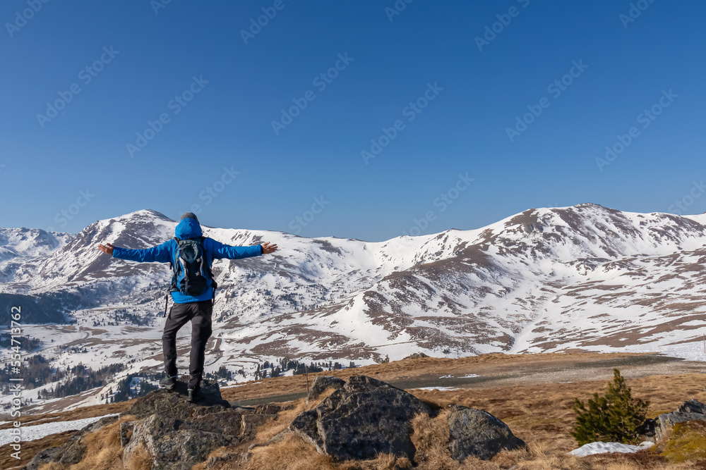 Rear view of man spreading arms with backpack looking at snow capped mountain peak Zirbitzkogel and Kreiskogel in Seetal Alps, Styria (Steiermark), Austria, Europe. Hiking trail in spring on sunny day