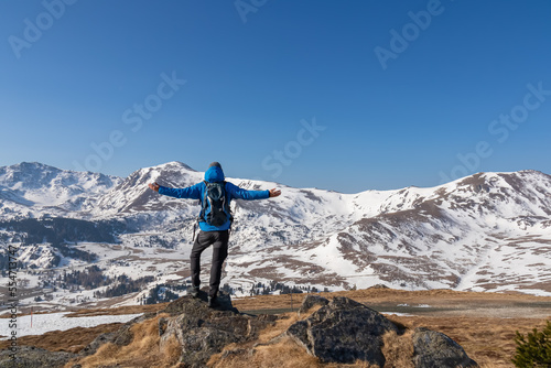 Rear view of man spreading arms with backpack looking at snow capped mountain peak Zirbitzkogel and Kreiskogel in Seetal Alps, Styria (Steiermark), Austria, Europe. Hiking trail in spring on sunny day