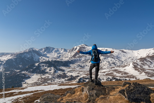 Rear view of man spreading arms with backpack looking at snow capped mountain peak Zirbitzkogel and Kreiskogel in Seetal Alps, Styria (Steiermark), Austria, Europe. Hiking trail in spring on sunny day