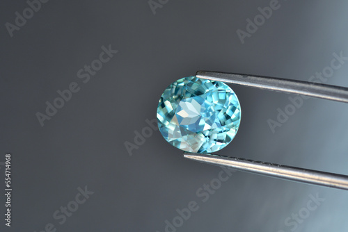 Genuine, natural mined, bright neon blue lagoon color, cushion shaped, heated loose zircon flawless gemstone setting for jewelry making. Light gray gradient background. Stone held in tweezers.