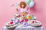 Surprised curly woman holds inflated balloons stands near messy table with variety of desserts has to bring house in order after birthday party wears festive dress isolated over pink background.