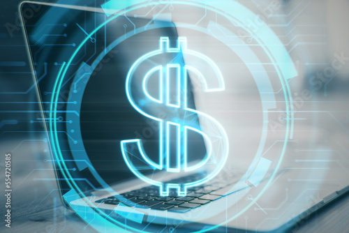 Close up of desktop with laptop and glowing dollar sign on blurry background. Money and online banking concept. Double exposure.