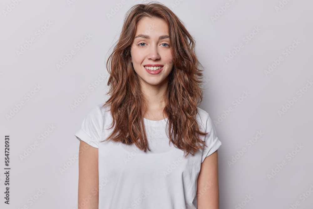 Portrait of beautiful blue eyed woman with dark wavy hair dressed in casual t shirt has natural beauty smiles toothily dressed in casual white t shirt isolated over greu background has healthy skin Portrait of beautiful blue eyed woman with dark wavy hair dressed in casual t shirt has natural beauty smiles toothily dressed in casual white t shirt isolated over greu background has healthy skin