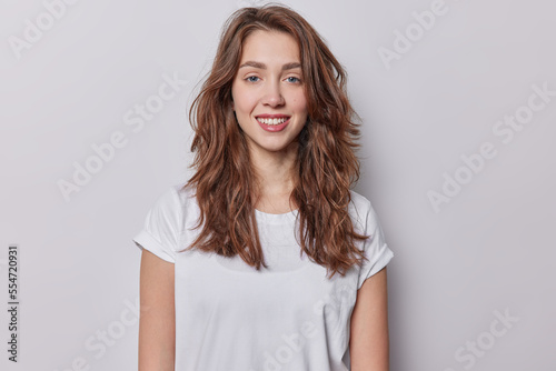 Portrait of beautiful blue eyed woman with dark wavy hair dressed in casual t shirt has natural beauty smiles toothily dressed in casual white t shirt isolated over greu background has healthy skin