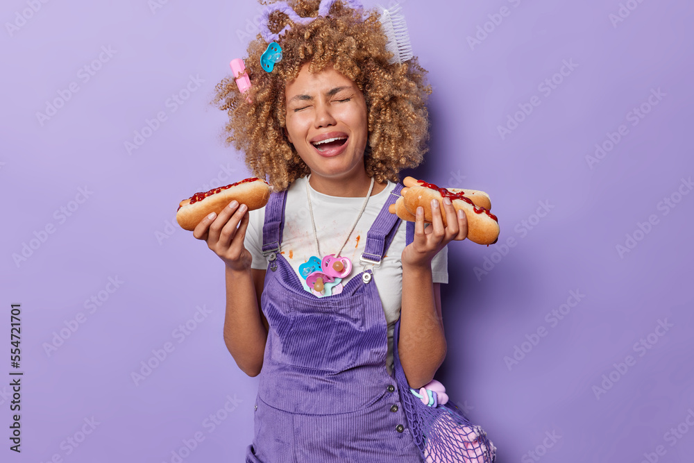 Displeased crying pregnant woman holds two hot dogs expresses negative emotions has addiction to unhealthy food dressed in casual clothes going to become mother soon isolated over purple background