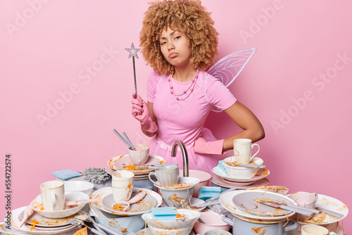 Obraz na plátně Serious self confident housewife with curly hair dressed like fairy holds magic wand going to make wish for doing washing up instead of you surrounded by pile of dirty dishes does housework