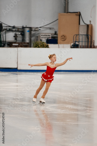 A little figure skater in a red and white tracksuit with a smile skates on ice at an indoor rink. Figure skating competition © Alena Vilgelm