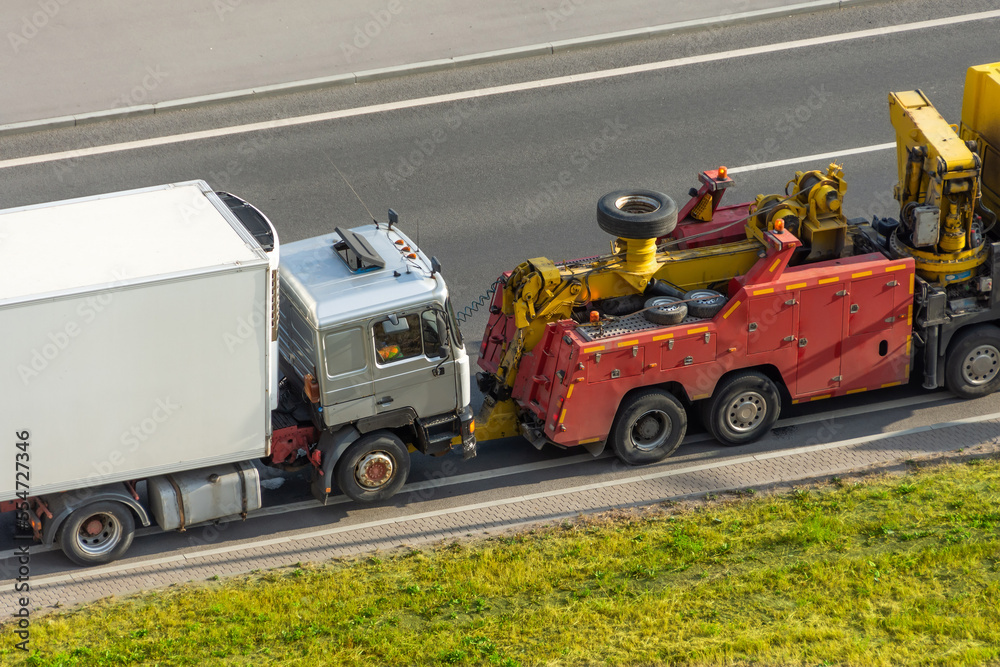 Powerful heavy duty big rig mobile tow semi truck with emergency lights and towing equipment prepare to tow broken white semi tractor standing out of service on the highway side.