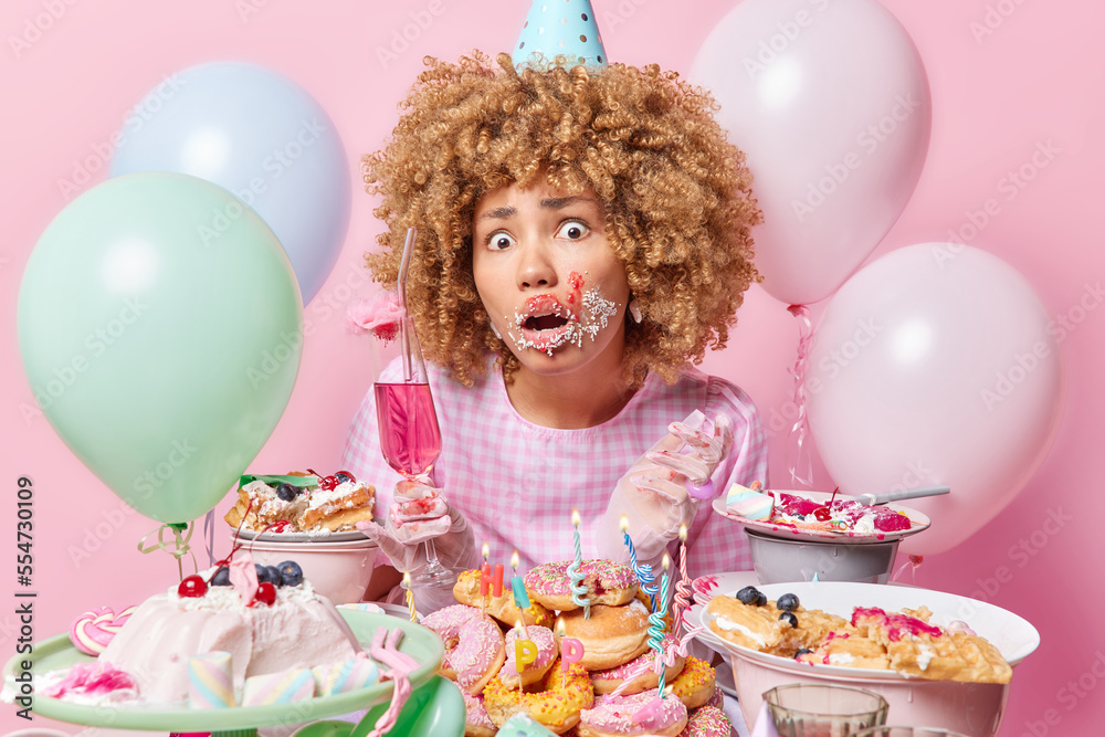 Impressed curly haired woman smeared with cream after eating cake surrounded by sweet doughnuts and jellies holds glass of cocktail wears party hat dress gloves poses around inflated balloons indoors Impressed curly haired woman smeared with cream after eating cake surrounded by sweet doughnuts and jellies holds glass of cocktail wears party hat dress gloves poses around inflated balloons indoors