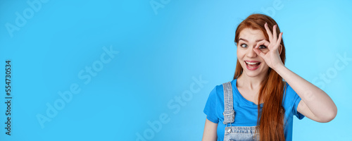 Cheerful upbeat redhead european female feel lucky inspired, look through circle okay on eye, smiling broadly amused, look wondered admire cool awesome sales, stand blue background