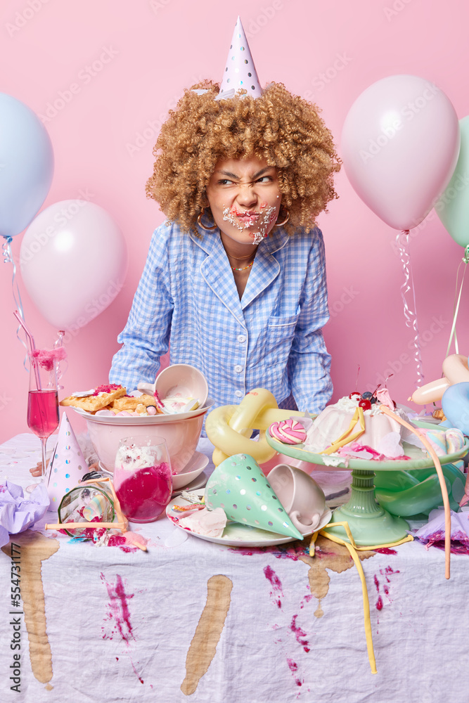 Displeased curly haired young woman wears cone hat and pajama stands ...