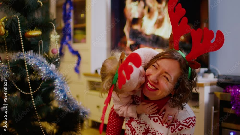 Happy caucasian mother hugging with daughter near the Christmas tree in a hat Happy caucasian mother hugging with daughter near the Christmas tree in a hat