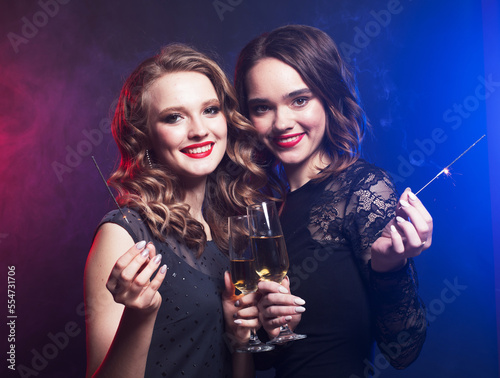 Celebration, party, and people concept - two young women in black cocktail dresses hold glasses of champagne.