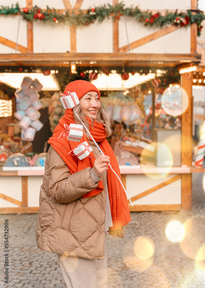 Asian woman in orange scarf and hat walking in Christmas market decorated with holiday lights. Feeling happy in  city Christmas decorated.