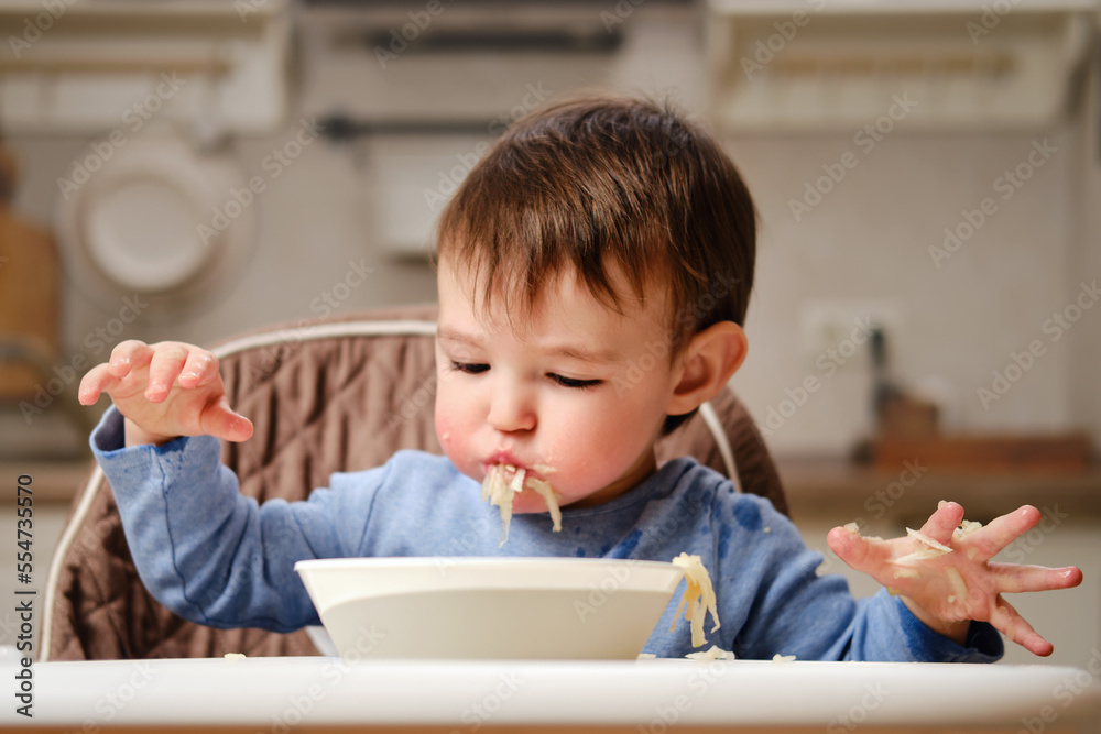 A funny child is eating a grated apple with his mouth full while sitting on a kitchen chair. Hungry baby boy shoves food in his mouth, humor. Kid aged one year four months A funny child is eating a grated apple with his mouth full while sitting on a kitchen chair. Hungry baby boy shoves food in his mouth, humor. Kid aged one year four months