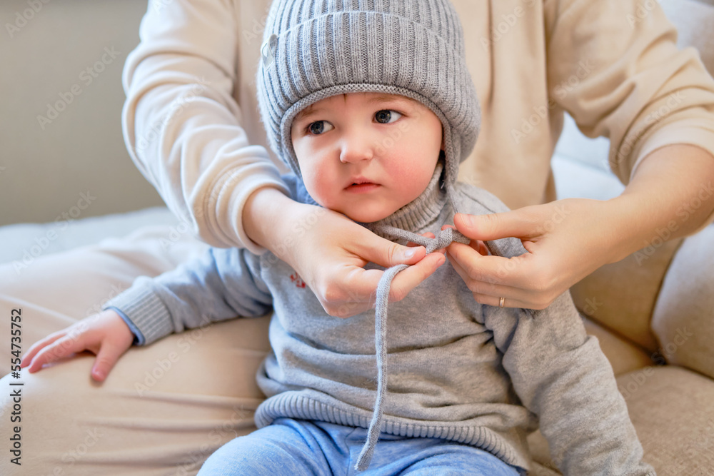 Child is dressed in warm winter clothes for a walk outside. Woman mother puts clothes on toddler baby boy for cold weather. Kid age one year five months in the home living room Child is dressed in warm winter clothes for a walk outside. Woman mother puts clothes on toddler baby boy for cold weather. Kid age one year five months in the home living room