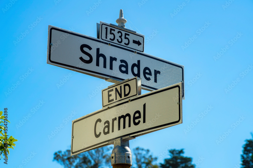 Sign that says Shrader and Carmel with white and black paint with foliage and trees in distance and clear blue sky background Sign that says Shrader and Carmel with white and black paint with foliage and trees in distance and clear blue sky background
