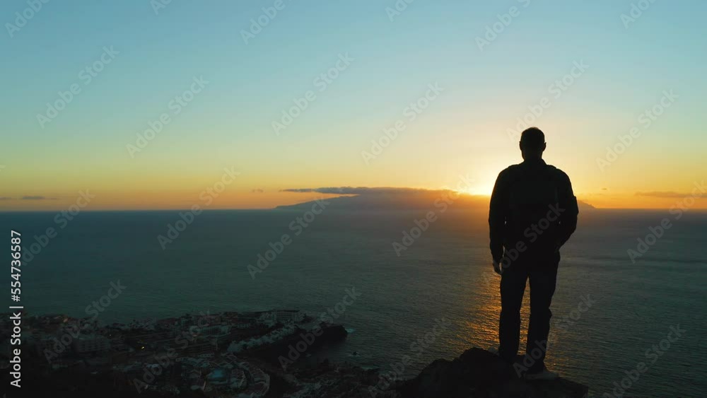 Silhouette of hiker man standing on mountain top at sunset. Boy looking at deep ocean and orange dark sky. Adventure freedom exploration of Planet Earth. Silhouette of hiker man standing on mountain top at sunset. Boy looking at deep ocean and orange dark sky. Adventure freedom exploration of Planet Earth.