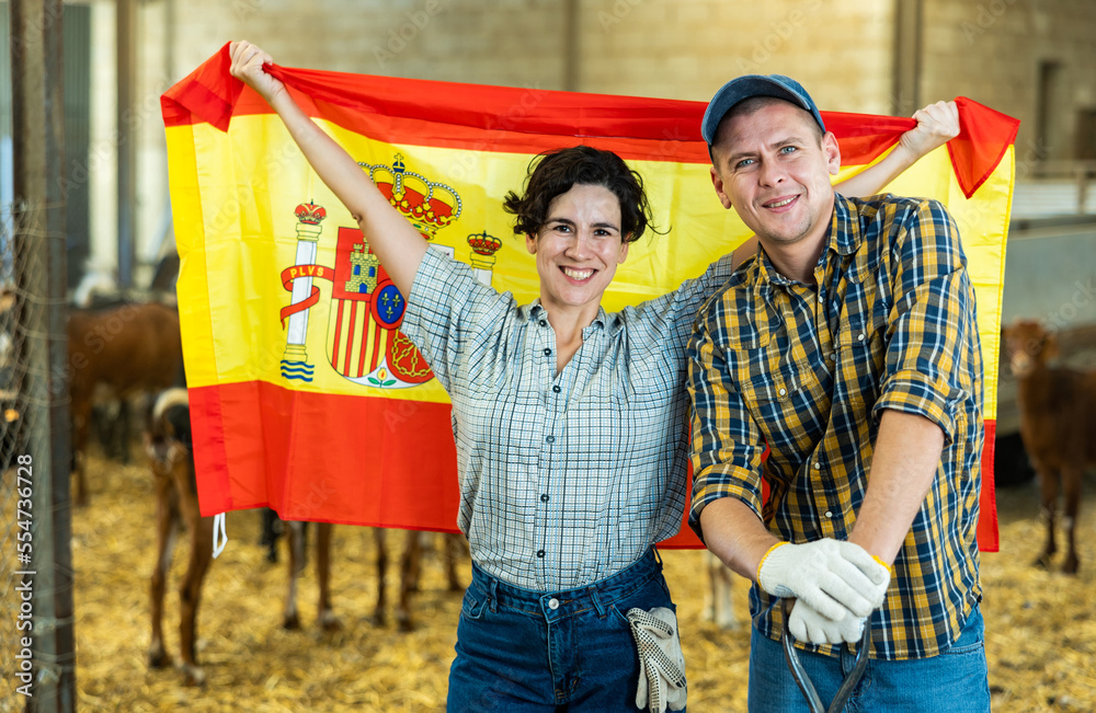 Excited young male and female farmer coworkers waving flag of Spain and smiling at camera on goat farm