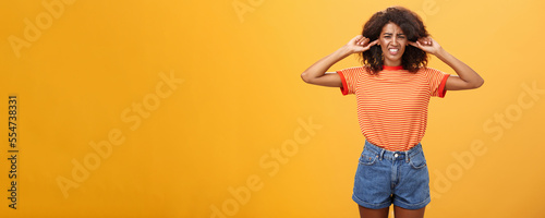 What that anoying sound. Disturbed and displeased dark-skinned girl with curly hair frowning clenching teeth from discomfort closing ears with index fingers from loud irritating sound over orange wall photo