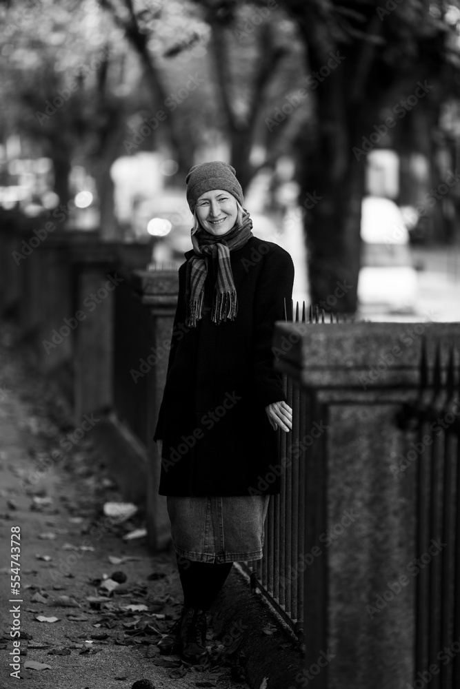 A happy woman in an autumn coat stands near a park fence. Black and white photo.