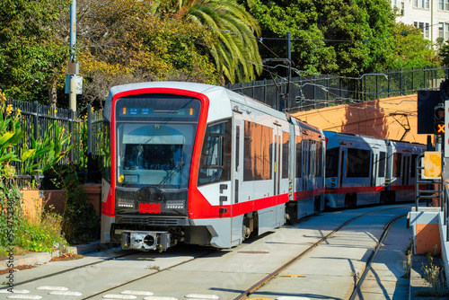 Tram or train with red and grey paint on railing with shrubs and trees in background in an urban part of the city downtown
