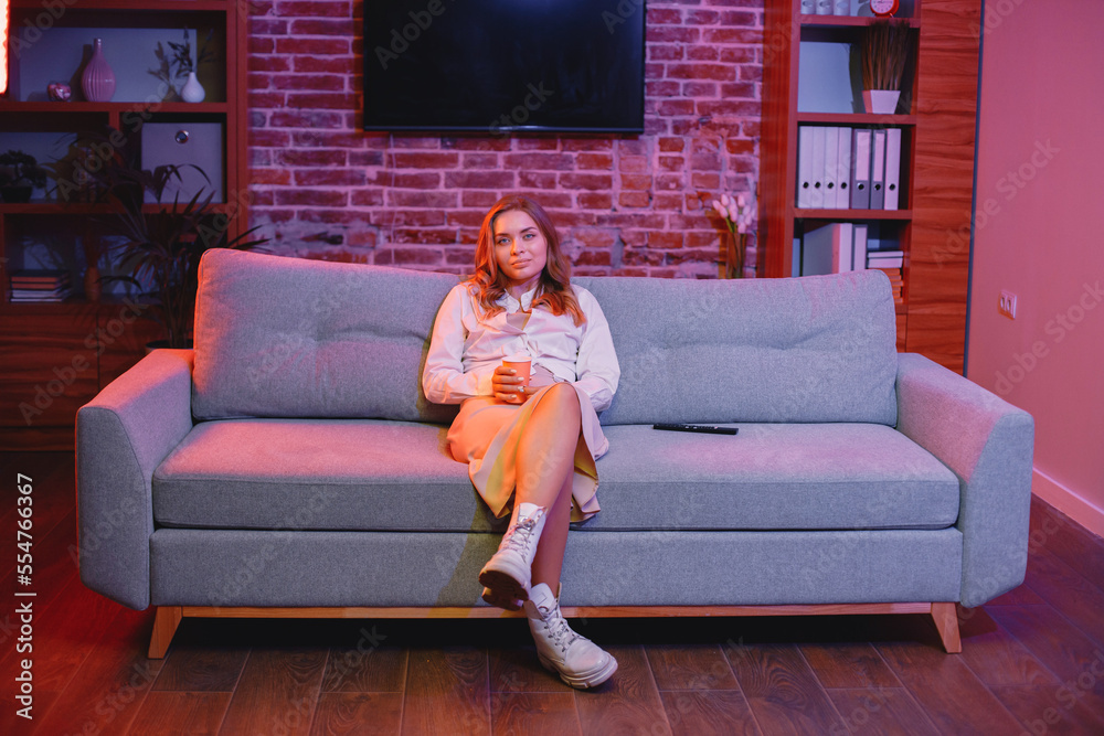 Obraz premium Young woman sitting on a sofa in a cozy room and looking directly at camera
