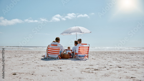 Family, relax and picnic in the sun on the beach for summer vacation, holiday or weekend getaway in the outdoors. People relaxing by the ocean coast with chairs and umbrella for free time in nature #554779756
