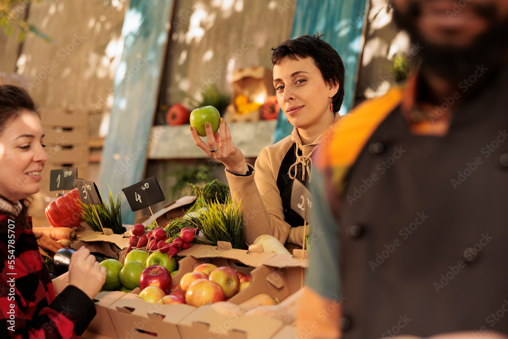Young female farmer showing various organic fruits to customers at local grocery marketplace. Woman working as vendor holding organic bio homegrown products, fresh seasonal food.