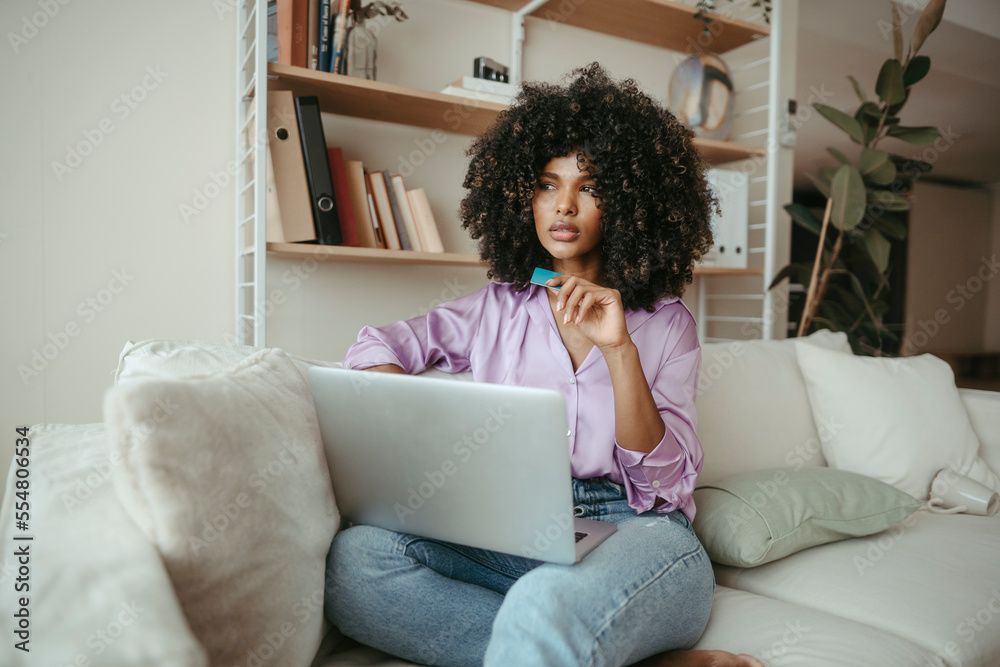Thoughtful woman with laptop and credit card sitting in living room Thoughtful woman with laptop and credit card sitting in living room