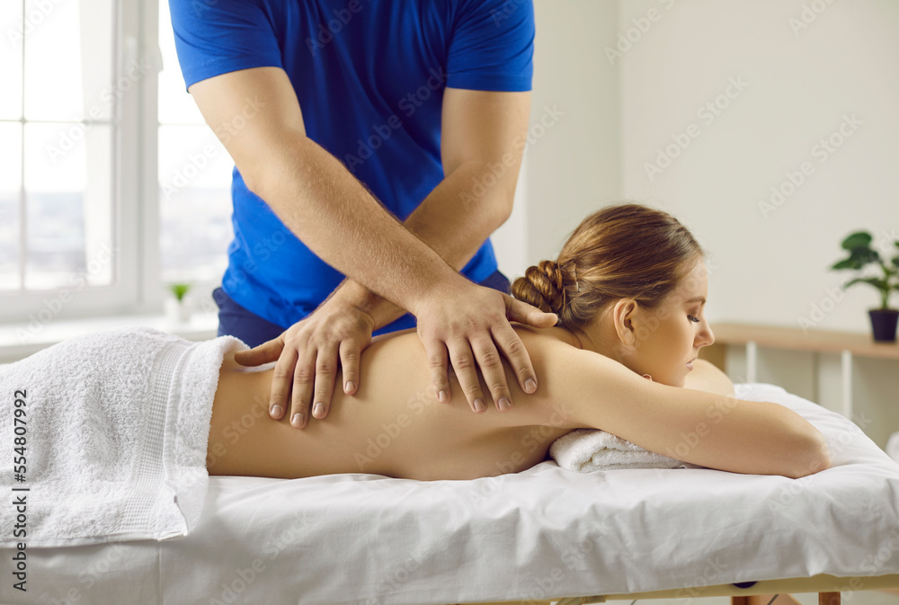Obraz premium Slender shirtless woman lying on her chest receives relaxing back massage in spa or massage parlor. Side view of male masseur massaging back of woman lying on massage table. Beauty concept.