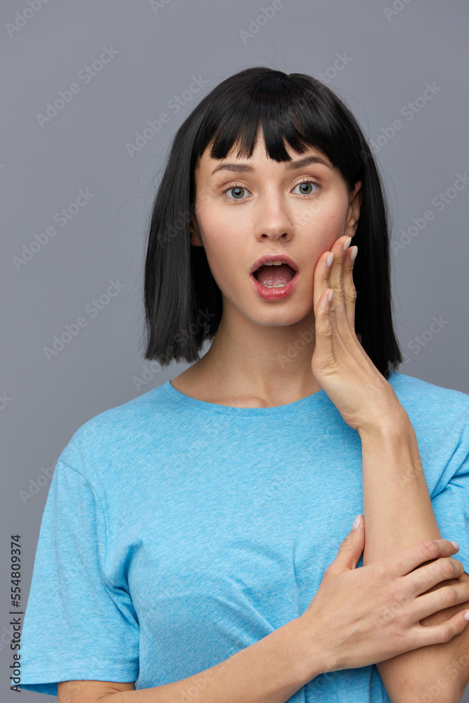 a pretty sweet woman standing on a gray background in a blue T-shirt opened her mouth in surprise, putting her hand to her cheek, holding her second a pretty sweet woman standing on a gray background in a blue T-shirt opened her mouth in surprise, putting her hand to her cheek, holding her second