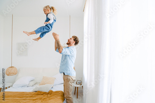 Fototapeta Naklejka Na Ścianę i Meble -  Young father throwing his little daughter in air and having fun at home