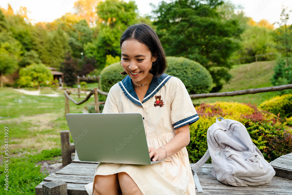 Cheerful asian woman using laptop while sitting on bench in green park Cheerful asian woman using laptop while sitting on bench in green park
