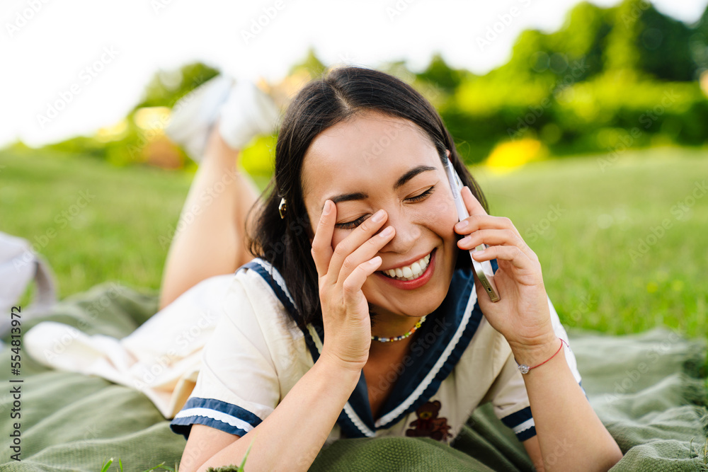 Cheerful asian woman talking on mobile phone while lying on blanket in park Cheerful asian woman talking on mobile phone while lying on blanket in park
