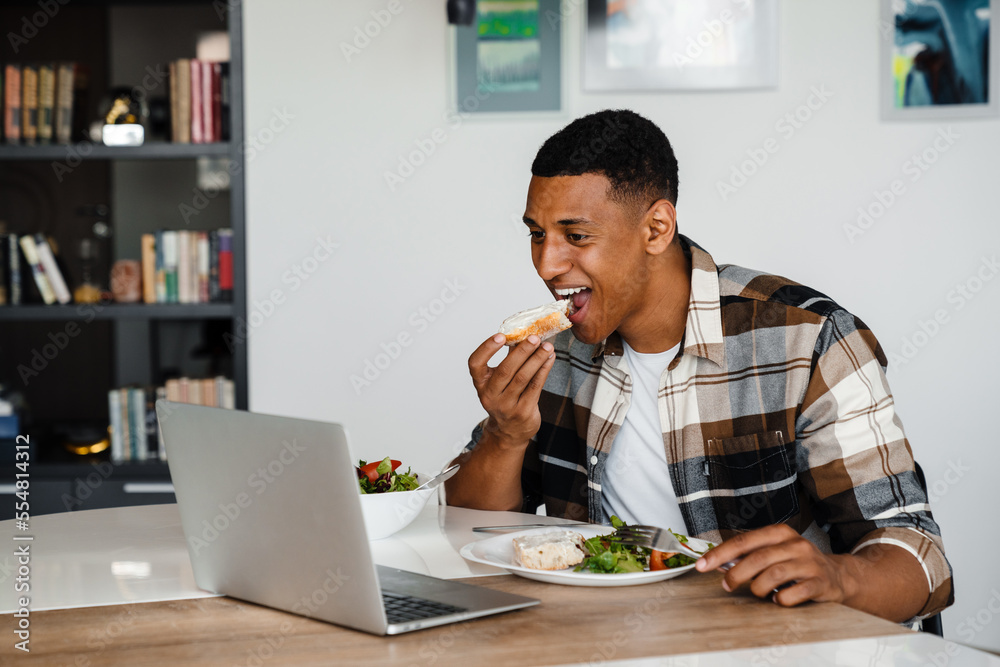 Young african man watching movie on laptop while having lunch at home Young african man watching movie on laptop while having lunch at home