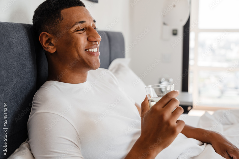 Young african man holding glass of water while lying in bed Young african man holding glass of water while lying in bed