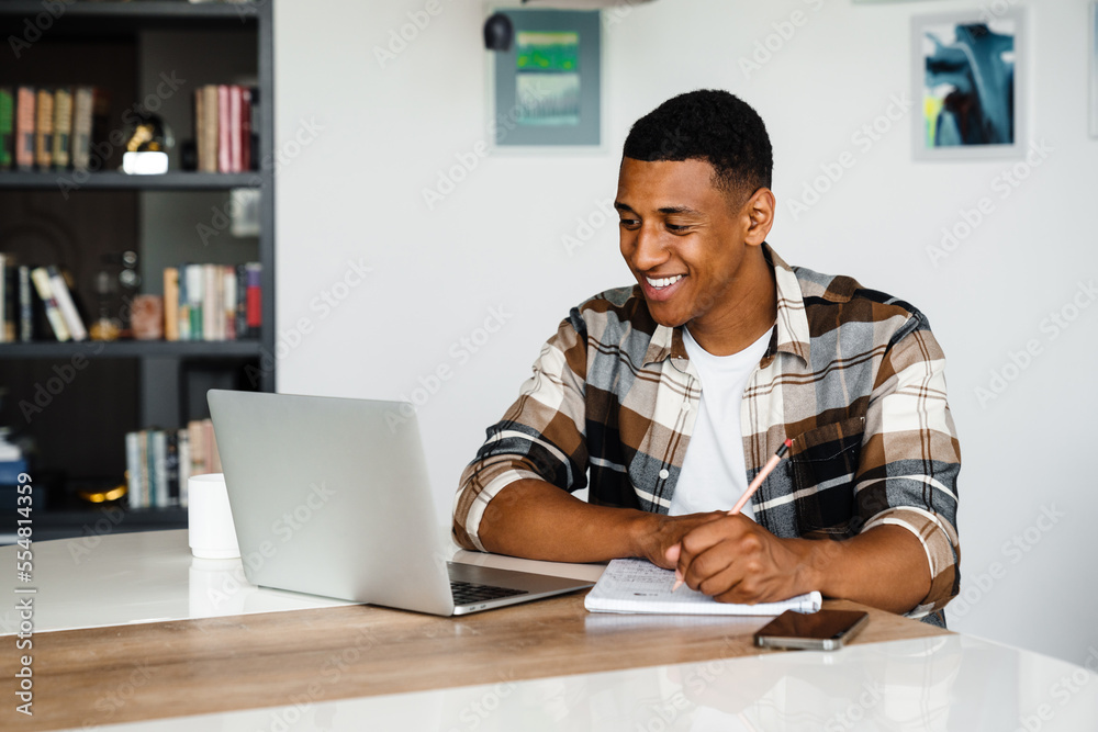 Young cheerful african man studying on laptop and writing down notes while sitting at table at home Young cheerful african man studying on laptop and writing down notes while sitting at table at home