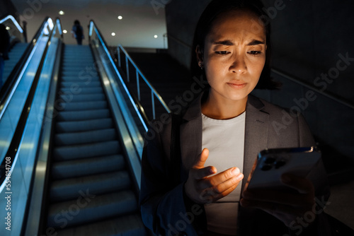 Fotografija Young asian woman frowning while using smartphone standing on escalator indoors