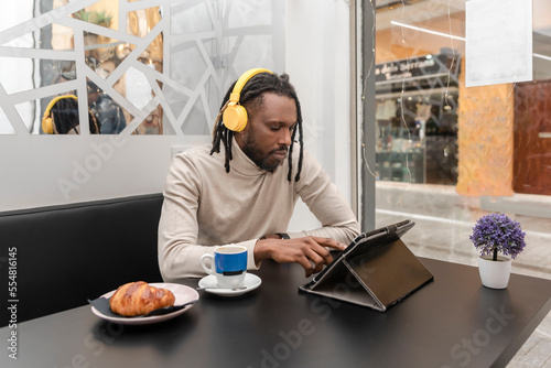 a modern man with dreadlocks in the cafeteria using a digital tablet while listening to music with yellow headphones