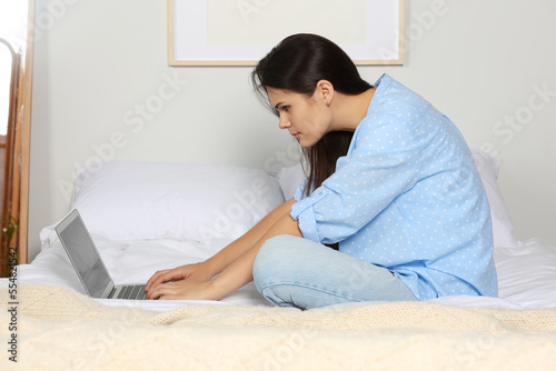 Young woman with bad posture using laptop while sitting on bed at home. Symptom of scoliosis