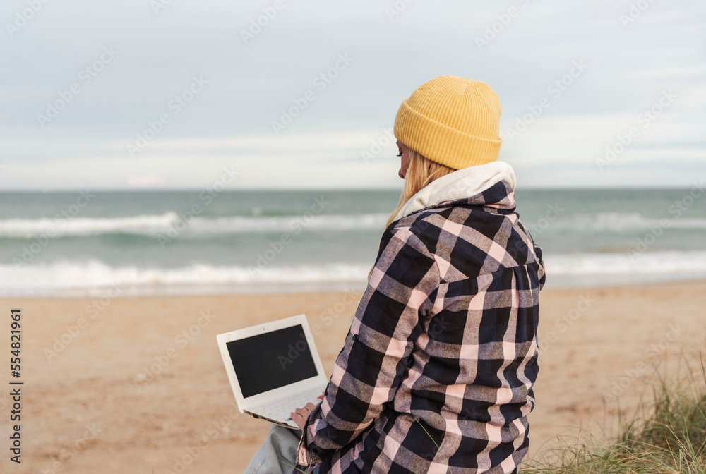 unrecognizable nomad digital woman in winter or autumn sitting on the beach using laptop