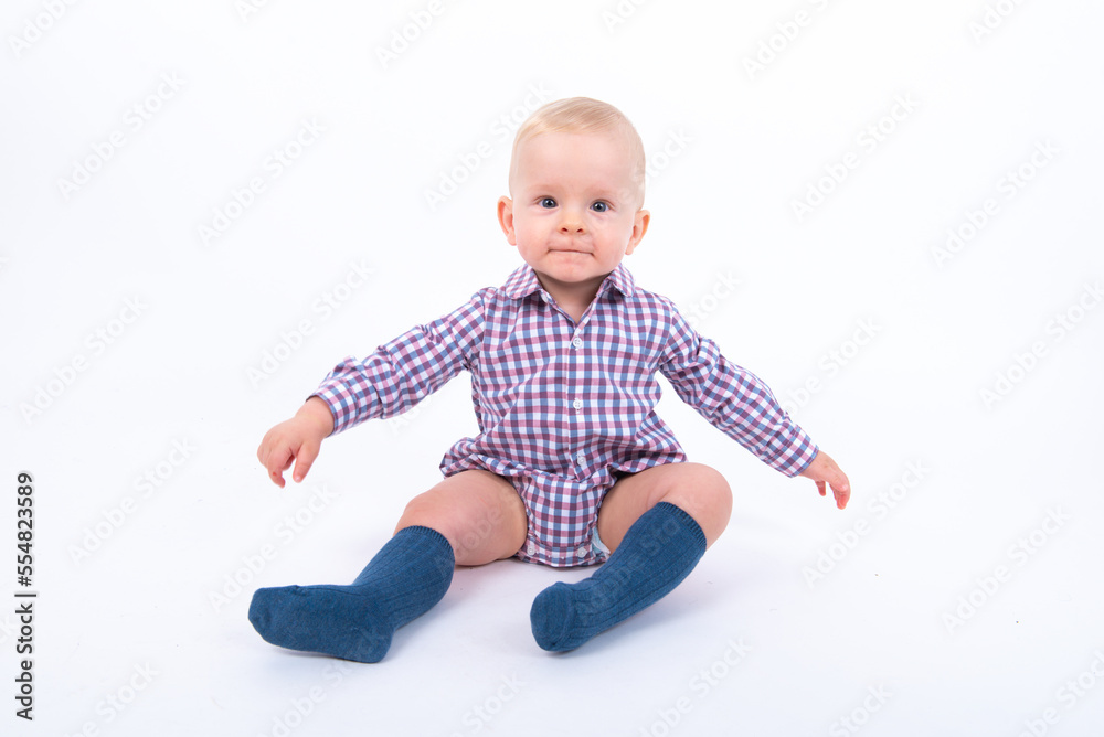 Playful beautiful baby boy wearing plaid bodysuit standing over white studio background sitting and smiling to the camera. Baby boy wants to play with his mother Playful beautiful baby boy wearing plaid bodysuit standing over white studio background sitting and smiling to the camera. Baby boy wants to play with his mother